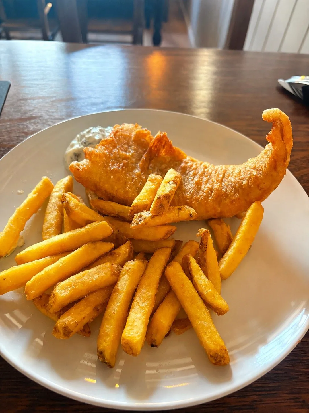 A plate of beer-battered cod with thick-cut chips and a pot of homemade tartare sauce, on a dark wooden table in the bar