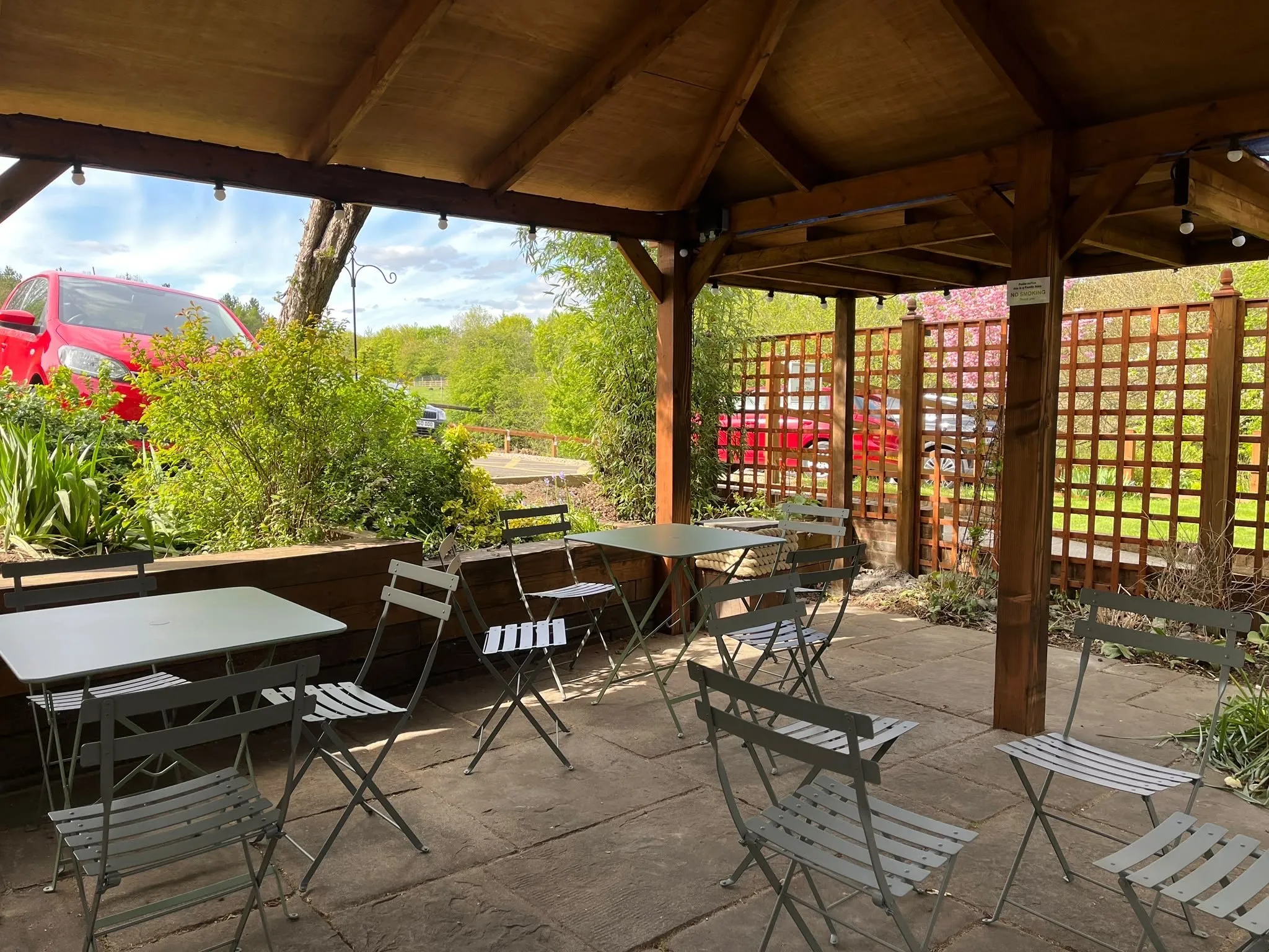 Seating under the covered pergola with festoon lights overhead, looking out past the trellis and garden beds towards the car park