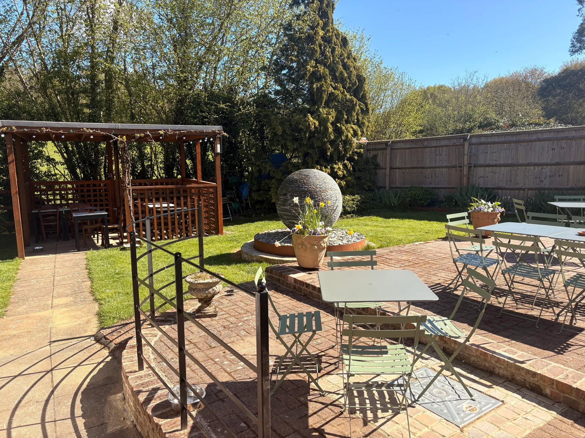 The Old Ship's sunny back garden: a timber pergola, a brick terrace with bistro tables, a stone sphere water feature and a wide lawn under blue sky
