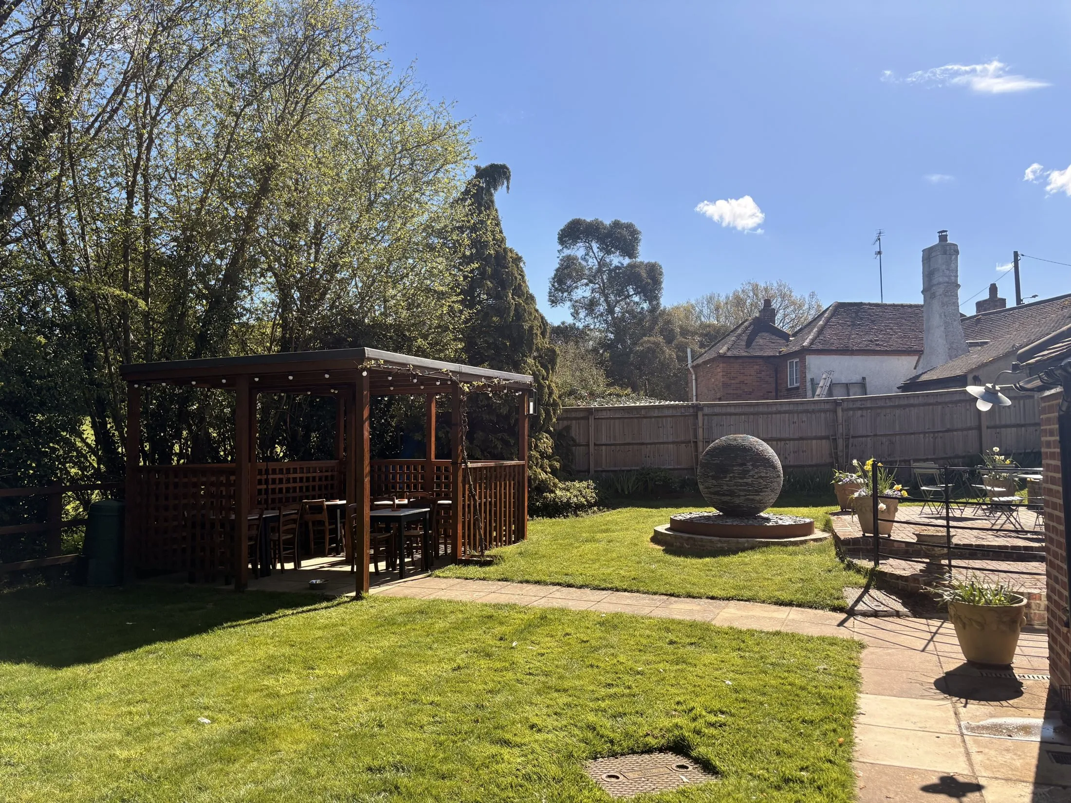A wide view of the garden: the timber pergola and its seating on the left, a stone sphere water feature, a lawn and terrace tables, with the pub beyond