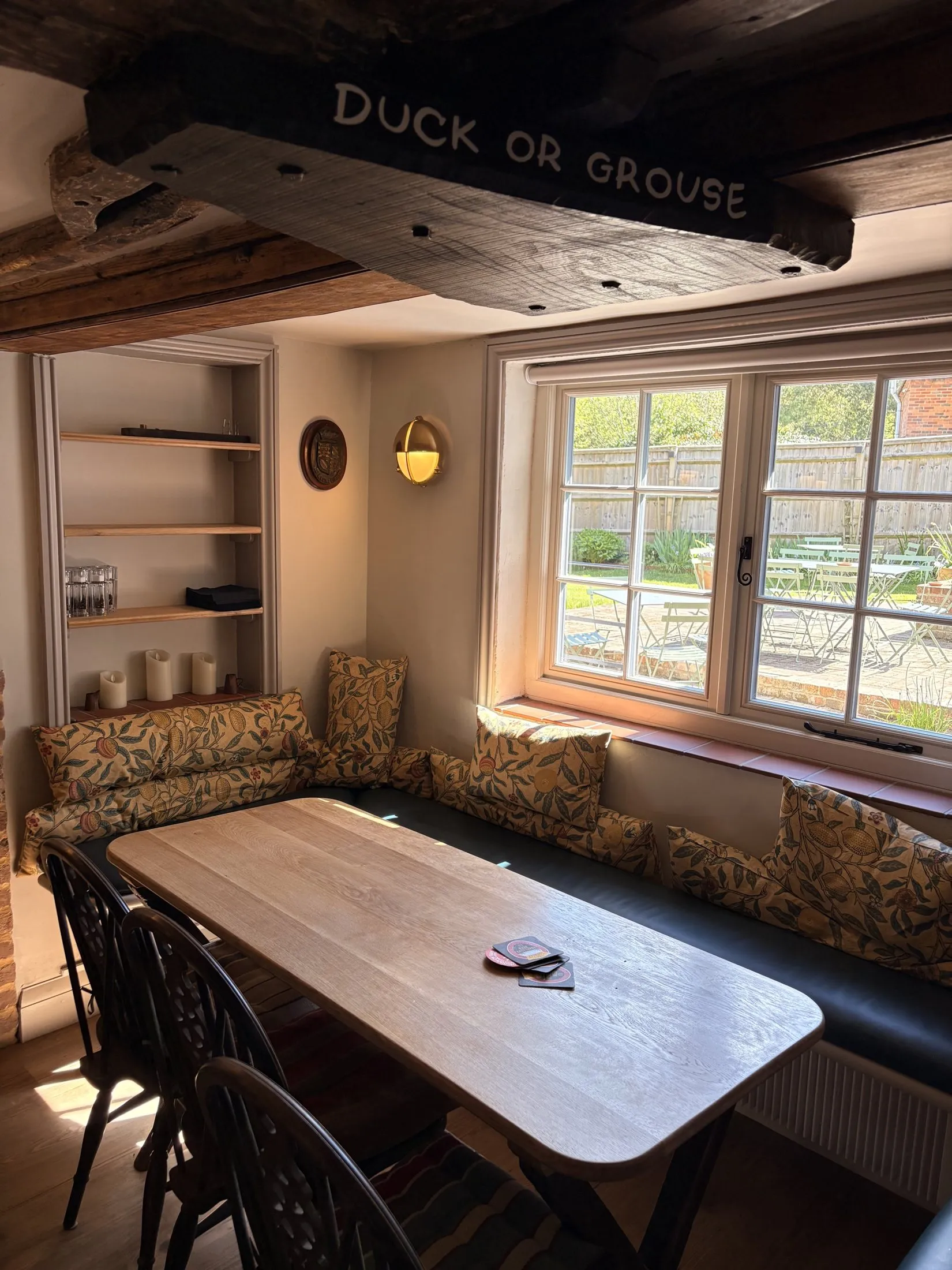 A snug corner of The Old Ship: a low beam painted 'Duck or Grouse', a cushioned window seat looking onto the garden, and a worn oak table set for a chat