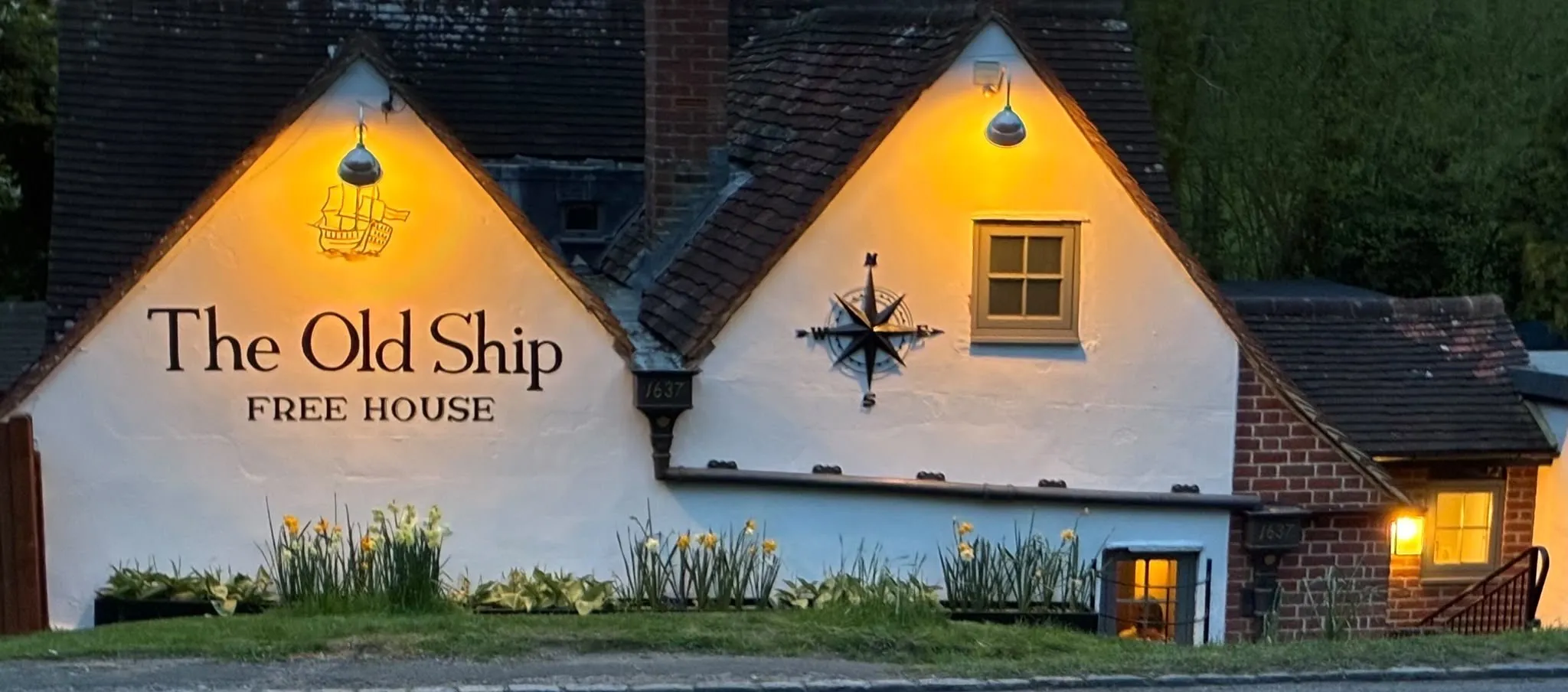 The Old Ship's whitewashed gables lit up at dusk, with 'The Old Ship, Free House' painted on the wall, a ship weathervane, the compass emblem beside a window, and daffodils along the front bed