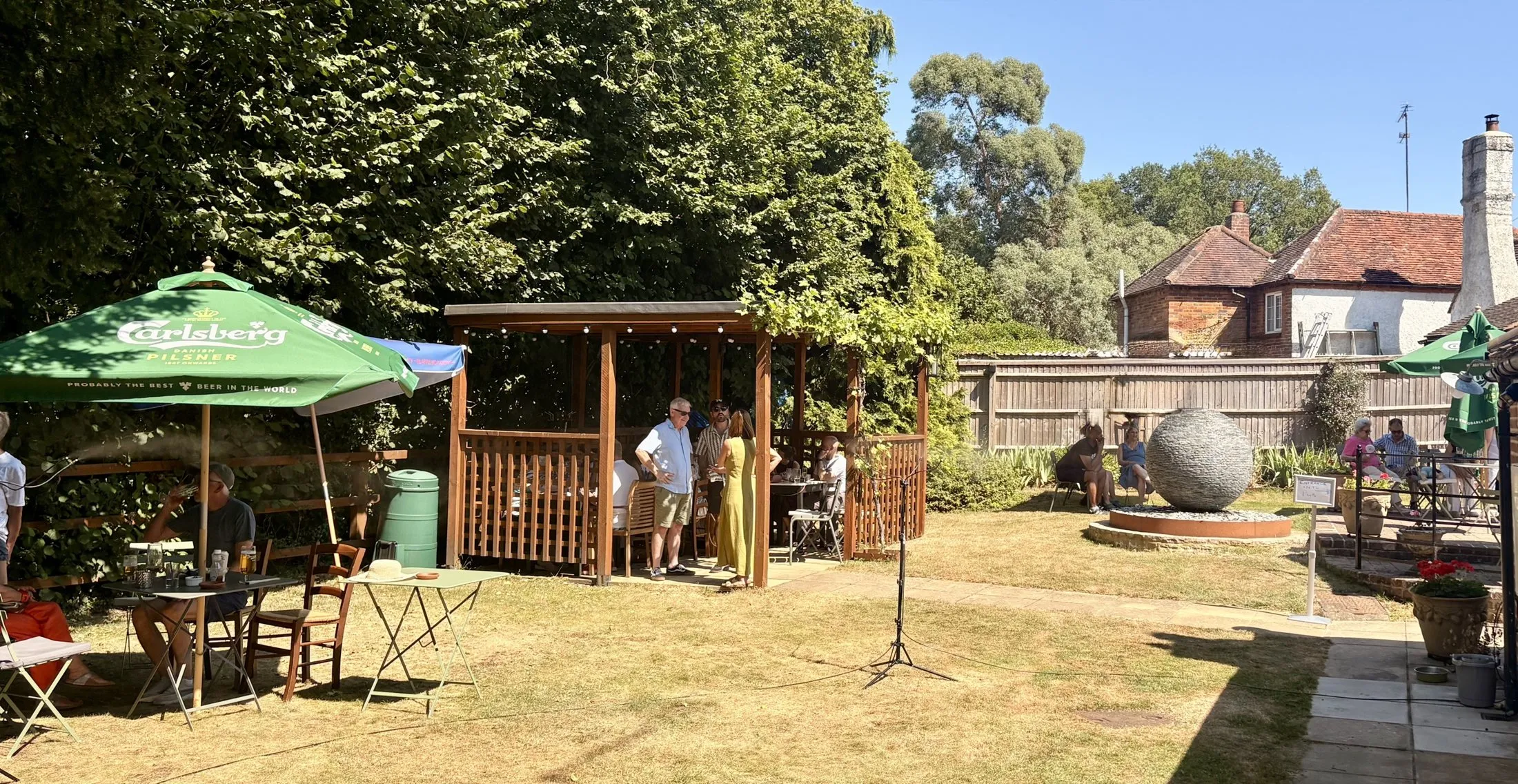 The Old Ship's garden on a sunny day: people gathered under the timber pergola, parasols and bistro tables on the lawn, a stone sphere water feature and the pub beyond